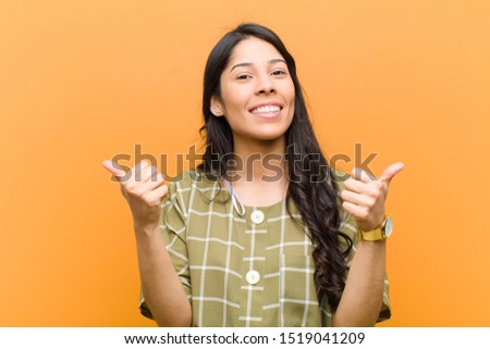 young pretty hispanic woman smiling joyfully and looking happy, feeling carefree and positive with both thumbs up against brown wall