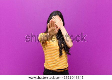 young pretty latin woman covering face with hand and putting other hand up front to stop camera, refusing photos or pictures against purple wall