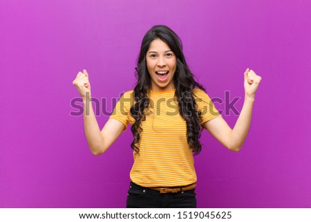 young pretty latin woman feeling happy, surprised and proud, shouting and celebrating success with a big smile against purple wall