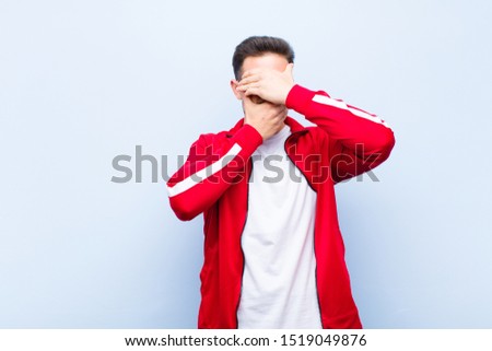 young handsome sports man or monitor covering face with both hands saying no to the camera! refusing pictures or forbidding photos against flat wall