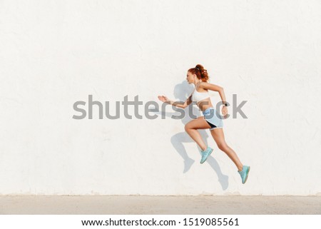 Image of redhead young woman in sportswear running along white wall while doing workout in morning