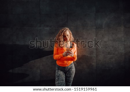 Charming smiling caucasian woman in sportswear leaning on black wall and using smart phone while resting after exercises.