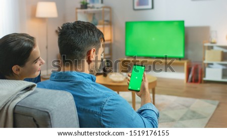 Happy Couple Sitting At Home in the Living Room Watching Green Chroma Key Screen Television, Relaxing on a Couch. Guy also Uses Green Mock-up Screen Smartphone.