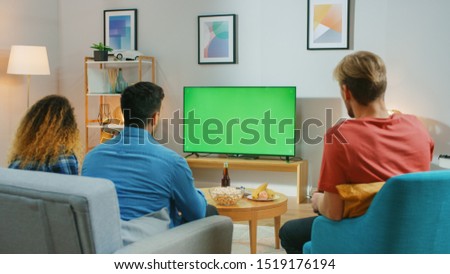 Diverse Group of Friends Sitting on a Couch at Home, Watch Green Chroma Key Screen TV while Eating Snacks and Drinking Beverage. Young People Having Fun at Home.