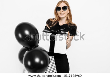 Happy young beautiful girl in black dress and sunglasses holding bags, balloons and gift box with black ribbon, on white background. Shopping, discounts, Black Friday