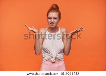 Horizontal indoor photo of pretty young woman with foxy hair combed in bun posing over orange background, shrugging perplexedly with raised hands and frowning her face