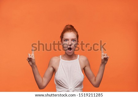 Indoor portrait of amazed attractive young redhead female with bun hairstyle wearing white top over orange background, rounding eyes with wide mouth opened and showing upwards with forefingers