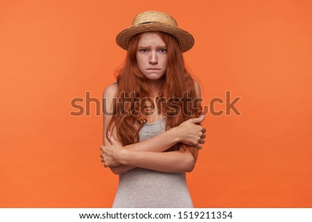 Studio portrait of frightened young beautiful redhead woman standing over orange background with folded hands, looking to camera scarely and wrinkling forehead