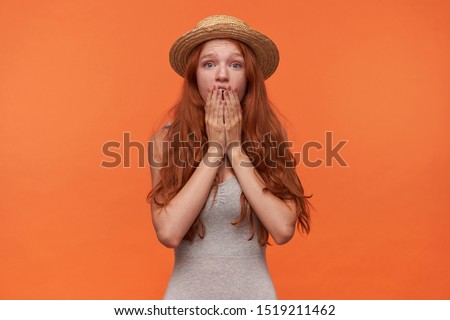 Horizontal shot of attractive young lond haired foxy female standing over orange background with shocked face, raising palms to her wide opened mouth, wearing grey shirt and straw hat