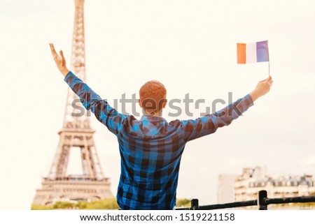 Smiling young man holding a French flag on the background of the famous Eiffel tower. Travel, immigration and study in Paris concept