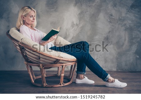 Full size profile side photo of concentrated middle aged woman sit in wicker armchair read paper book love story on autumn holidays in  room indoors
