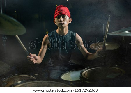 Teenager rock band drummer . 13 or 14 years old cool and talented Asian American mixed ethnicity young boy playing drums in headband performing song in dark stage background