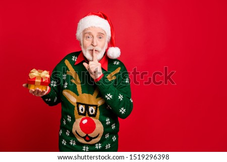 Hush secret! Portrait of funky grey hair old man in santa claus headwear hold little package show mute sign say dont tell prepare x-mas present wear antler decor sweater isolated red color background