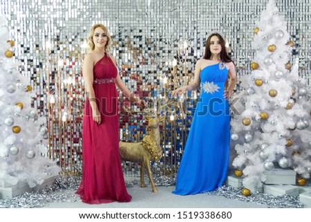 Beautiful young girls wearing red and blue dresses posing on the New Year decoration background with white decorated Christmas trees. Festive Christmas party