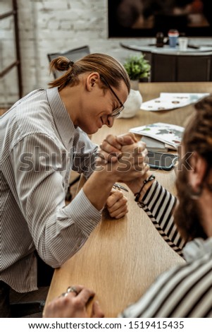 Do your best. Cheerful male person pressing his eyes while doing arm-wrestling with his colleague