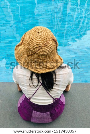 Asian woman tourist sit back and wearing the  woven hat sit by the blue swimming pool side in hotel to relax during the holidays vacation on summer season 