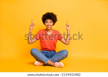 Full length body size photo of cute pretty sweet curly girl pointing up to show you sales sitting on floor with legs crossed wearing orange t-shirt jeans denim isolated vivid color background