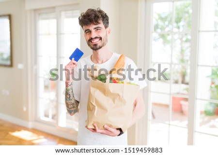 Young handsome man holding a paper bag full of fresh groceries at home, showing credit card as payment metod