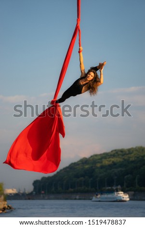 Beautiful and flexible female circus artist dancing with aerial silk with sky and river coastline on background