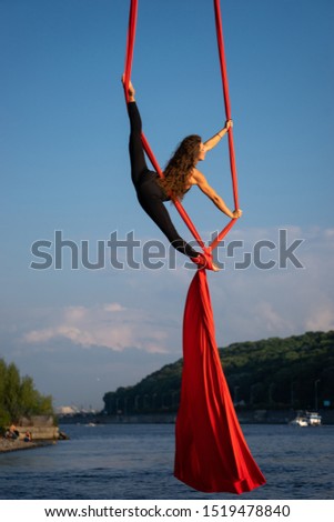 Beautiful and flexible female circus artist dancing with aerial silk with sky and river coastline on background