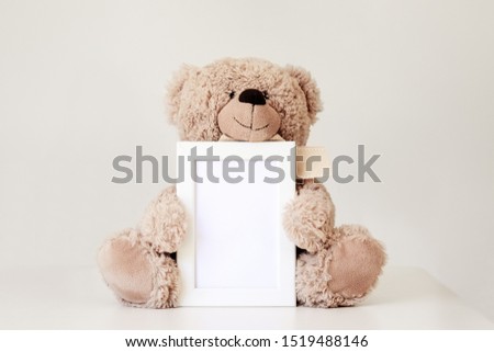 Mockup. Soft beige teddy bear toy holding white clean mock up frame with copy space sitting at light grey background. Empty space. Baby children concept. 