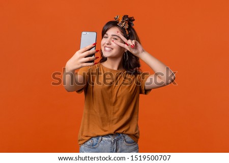 Pretty brunette woman in a t-shirt and beautiful headband making selfie with spartphone and sending air kisses standing isolated over orange background. Mobile photography