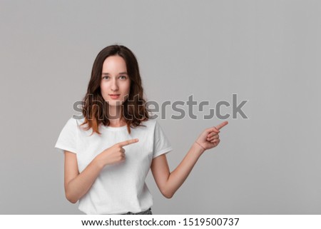 Pretty brunette woman in a white tee pointing at the empty space in a right side of a frame standing isolated over grey background. Copy space of advertising.