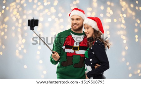 christmas, technology and holidays concept - happy couple in santa hats taking picture by smartphone on selfie stick at ugly sweater party over festive lights background