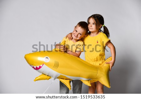 Little girl in a yellow dress hugs her brother friend and holds a balloon in the form of a shark in the studio on a gray background