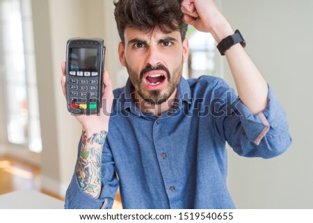 Young man holding dataphone point of sale as payment annoyed and frustrated shouting with anger, crazy and yelling with raised hand, anger concept