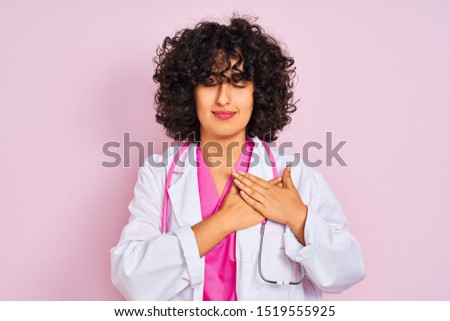 Young arab doctor woman with curly hair wearing stethoscope over isolated pink background smiling with hands on chest with closed eyes and grateful gesture on face. Health concept.