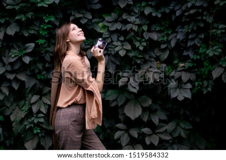 young beautiful girl photographer stands with a film camera near a wall of leaves in the forest, a woman photographs in nature