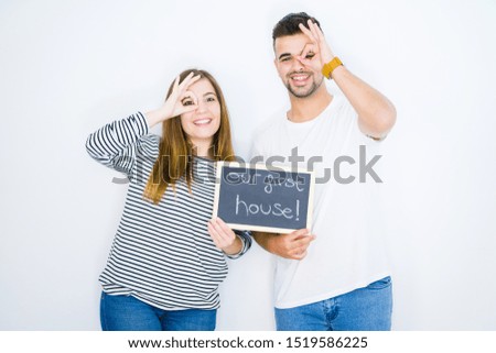 Young couple holding blackboard with our first home text over white isolated background with happy face smiling doing ok sign with hand on eye looking through fingers