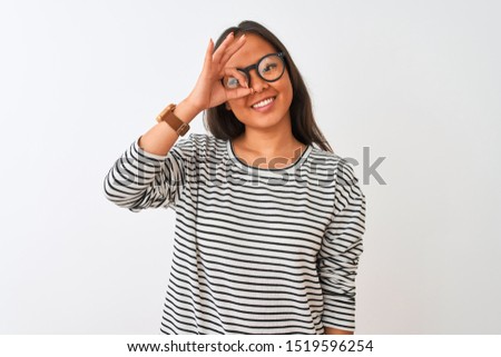 Young chinese woman wearing striped t-shirt and glasses over isolated white background doing ok gesture with hand smiling, eye looking through fingers with happy face.