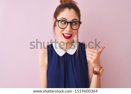 Redhead woman with pigtail wearing elegant dress and glasses over isolated pink background smiling with happy face looking and pointing to the side with thumb up.