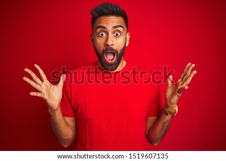 Young handsome indian man wearing t-shirt over isolated red background celebrating crazy and amazed for success with arms raised and open eyes screaming excited. Winner concept