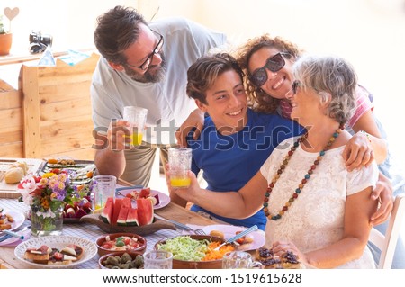 Cheerful family, three generations, hugging each other eating vegan food together on the wooden table. Senior woman with son, daughter-in-law and nephew. Bright sunlight.