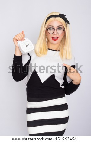 Beautiful blonde woman in glasses holding and looking at alarm clock in her hands