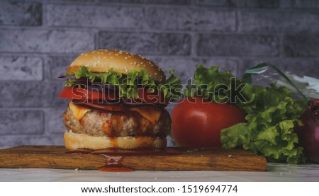 A huge juicy hamburger is on a chopping board with vegetables on a gray brick background.