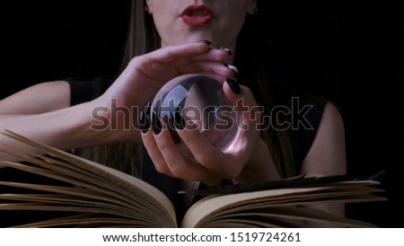 Attractive mystic witch woman with bright red lips on a black background, looks into a magic ball while casting spells next to a spell book. front view