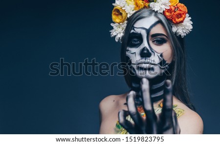 Young female in floral wreath and with spooky skeleton makeup gesticulating and looking at camera while luring prey during Halloween party against dark blue background