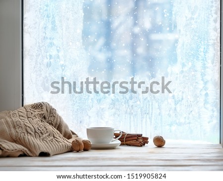 frozen window and cup of tea or coffee, sweater, cinnamon sticks, nuts on windowsill. cozy mood, home comfort in snowy cold weather. festive winter season. Christmas, New Year holidays background.
