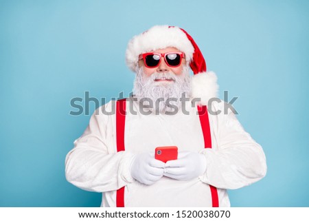 Close up photo of funny funky fat white grey-haired santa claus use cell phone type sms congratulations with noel in blogs wear overalls  red suspenders sunglass isolated over blue color background