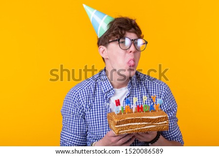 Crazy cheerful young man in paper congratulatory hat holding cakes happy birthday standing on a yellow background. Jubilee congratulations concept.