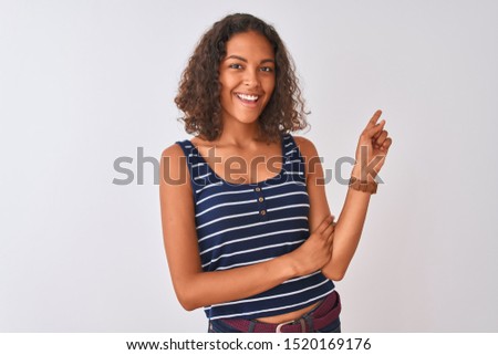 Young brazilian woman wearing striped t-shirt standing over isolated white background with a big smile on face, pointing with hand and finger to the side looking at the camera.