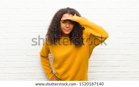 young black woman looking bewildered and astonished, with hand over forehead looking far away, watching or searching against brick wall