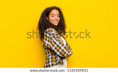 young pretty african american woman smiling to camera with crossed arms and a happy, confident, satisfied expression, lateral view against yellow wall