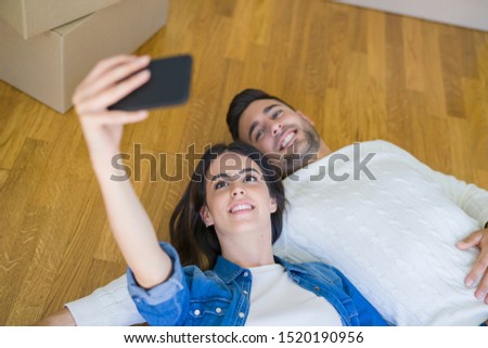 Young couple lying on the floor of new house taking a selfie photo using smartphone, smiling happy for moving to new apartment