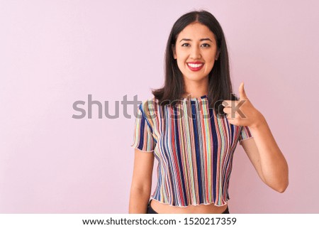 Young chinese woman wearing colorful striped t-shirt standing over isolated pink background doing happy thumbs up gesture with hand. Approving expression looking at the camera with showing success.