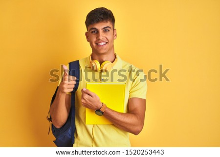 Indian student man wearing backpack headphones notebook over isolated yellow background happy with big smile doing ok sign, thumb up with fingers, excellent sign